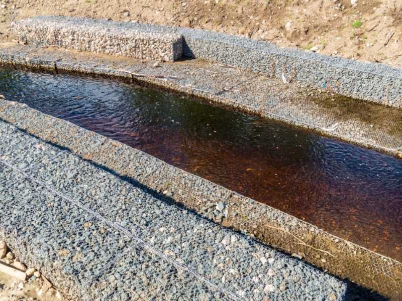 Gabion retaining wall erosion control | Frisco Civil Engineering A narrow concrete water channel or trench with dark water, bordered by rough gravel walls and soil beyond.
